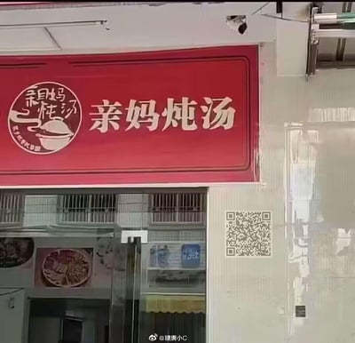 这是一家主打炖汤的线下餐饮店铺的门头招牌，整体为红色底色搭配白色字体，视觉醒目。招牌左侧是圆形品牌logo，内有“亲妈炖汤”的艺术字体、汤碗图案以及小字“良心炖汤好味道”；右侧是大号的“亲妈炖汤”字样，传递出像家人炖煮的暖心家常感。招牌下方是店铺的玻璃门，门内可见餐品海报，门外墙面贴有二维码，背景能看到带防盗窗的居民楼建筑。