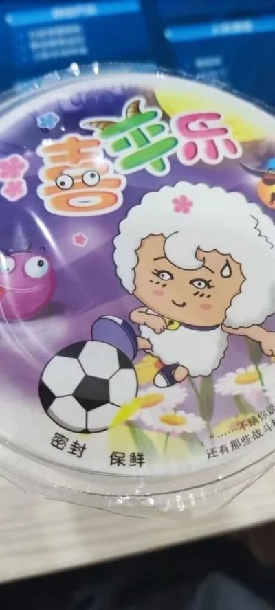 这是一张食品密封包装的实拍图，包装主色调为紫色，整体被透明塑料膜包裹。包装顶部印有彩色的“喜苹乐”字样，是对知名动画《喜羊羊与灰太狼》中“喜羊羊”的谐音改编。包装中间是模仿该动画中美羊羊的卡通形象：白色卷毛搭配粉色小花发饰，脸上挂着汗珠，正做出踢足球的动作，脚边是一颗黑白相间的足球。包装左侧还有一个粉色的卡通小怪物形象，睁着大眼睛，表情呆萌。包装底部印有白色的“密封 保鲜”字样，右下角则有一段被截断的文字“……不瞒你说 还有那些战斗”。该包装明显借助知名动画IP的谐音和形象进行模仿，以此来吸引消费者，尤其是儿童群体。
