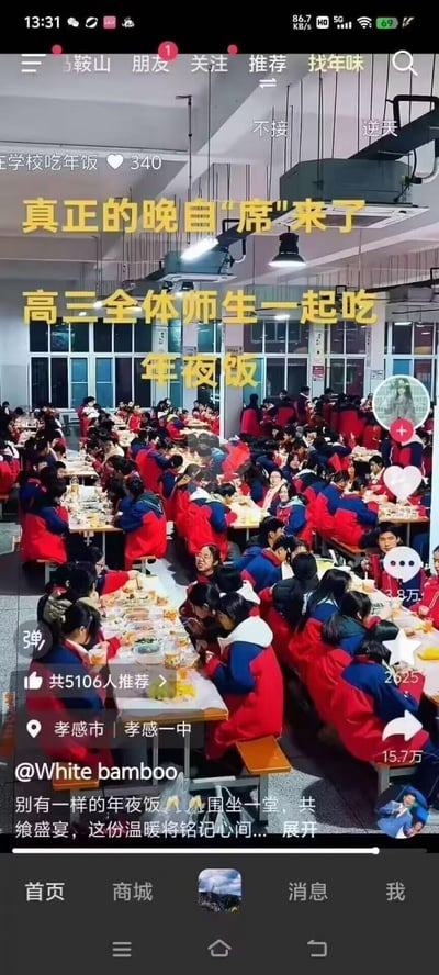 这是一张抖音平台的短视频截图，画面展示了孝感市孝感一中的高三全体师生在学校食堂围坐在一起同吃年夜饭的场景，学生们统一穿着红蓝相间的校服，餐桌上摆满了丰盛的餐食。图中的文字“真正的晚自‘席’来了”运用了谐音梗，将“晚自习”谐音为“晚自席”，既调侃了高三学生特殊的过年状态，也传递出留校过年的温暖氛围。视频发布者配文描述了这份别样年夜饭的暖心，称这份温暖会被铭记心间，该视频在抖音获得了大量用户的推荐和关注。