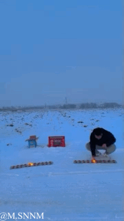 这是一段雪地场景的动态视频，画面里一名男子在雪地上摆放了两串类似鞭炮的长条状物品，他蹲下身点燃自己面前的一串，结果刚点燃就炸到了自己，而另一串毫无反应，场面充满反差感，十分搞笑。