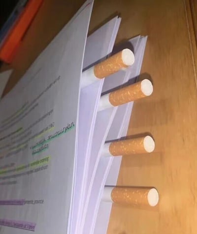 一张办公桌场景照片，展示了几支香烟被垂直插在一叠文件纸张之间，充当临时书签使用。文件上可见部分彩色文字，但内容不清晰。这种非常规的书签用法形成了画面的主要视觉焦点。