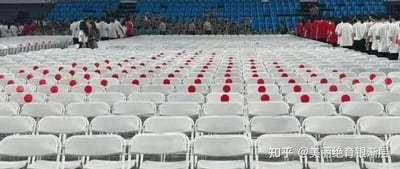 这是一张大型场馆内的实拍照片，场馆中整齐排列着大量白色折叠座椅，许多座椅的靠背上都被贴上了红色圆形贴纸。从拍摄的视角来看，这些红色圆点与白色椅背组合后，形成了类似乳腺的视觉错位效果，带来趣味的视觉反差。场馆后方聚集着不少身着白色和红色上衣的人员，看起来像是活动现场的工作人员或参与者，似乎在进行活动前的准备工作。