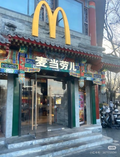 图片展示了一家融合传统中式建筑风格的麦当劳门店。建筑顶部采用典型的中式歇山顶设计，覆盖灰色瓦片，屋檐下装饰有色彩鲜艳的斗拱和彩绘纹样，包含红、绿、蓝等传统配色。正中央上方悬挂着麦当劳标志性的金色拱门logo，下方门楣处有“麦当劳儿”的中文招牌。门店入口为玻璃门，门两侧有绿色立柱，门前台阶上停放着几辆摩托车，背景可见光秃秃的树木和晴朗的天空，推测为冬季或初春白天场景。