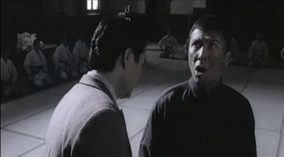 黑白影视截图中，两位男性在传统道场中央正面对峙。右侧男子（叶问）身着深色短衫，表情激动，嘴巴微张似在说话；左侧男子穿浅色长衫，身体微微前倾，神情严肃。背景中可见多名身着白色武道服的人整齐跪坐，注视着中央对峙场面，整体画面呈现出紧张的武术对决前氛围。