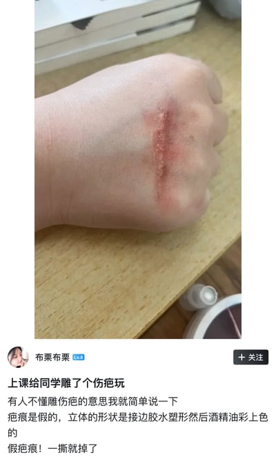 这是一张竖屏格式的社交媒体帖子截图，图片上半部分是实拍内容：一只握成拳的真人手放置在木质桌面上，手背上制作了一道长条形立体仿真假伤疤，伤疤效果逼真，视觉上类似真实的血肉伤口；图片下半部分是该帖子发布者的文字说明，发布者网名为布栗布栗，说明这个假伤疤是上课的时候给同学制作用来玩耍的，介绍了制作方法是用接边胶水塑形之后用酒精油彩上色，强调这是可以直接撕掉的假疤痕。