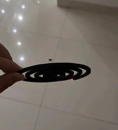 这是一张实拍的生活趣味照片，背景为室内光滑的浅色瓷砖地面，地面反射着顶部的点状灯光。画面左侧伸出一只手，用两根手指捏着一盘黑色螺旋结构的盘式蚊香，蚊香的一端已经点燃，露出红色的燃烧点。反常的是，有一只蚊子正平稳地停在蚊香的中心区域，没有受到蚊香驱蚊效果的影响，画面充满反差的幽默感。
