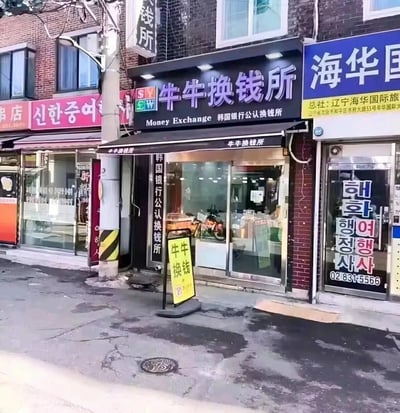 图片展示了一条街道的店铺门面场景，主体是一家名为“牛牛换钱所”的货币兑换机构。其招牌为黑色底，紫色和白色文字，上方标有“牛牛换钱所”，下方有“Money Exchange 韩国银行公认换钱所”的中英文说明，门口还立着一个黄色的“牛牛换钱”指示牌。左侧有红色招牌的店铺，标有“신한중약”（韩文），右侧是蓝色招牌的“海华国际”，显示总社位于辽宁。背景为砖结构建筑，街道地面有裂缝和井盖，整体呈现真实的城市街景风貌。