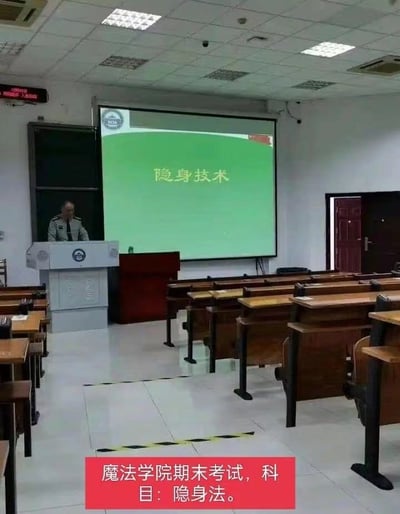 这张图拍摄的是普通高校的阶梯教室场景，讲台上站有一位身着制服的男性站在讲台后方，前方的投影幕布上显示着课程标题“隐身技术”，教室的学生座位区域全部空置没有任何学生在场；图片底部额外添加了红色背景的白色文字注释“魔法学院期末考试，科目：隐身法。”，利用空教室的场景配合投影的“隐身技术”课程名进行调侃，将现实中的理工科课程戏谑为魔法学院的隐身术考试，暗指在场的考生都已经成功使用了隐身法术，因此教室里看不到人，形成了幽默的玩梗效果。