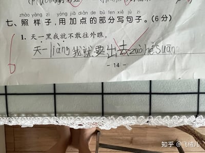 图片展示了一张小学生语文作业纸，上面是“照样子写句子”的练习。题目要求用“一...就...”结构造句，例句为“天一黑我就不敢往外瞧”。学生则写下“天一liàng我就要出去zuò hé suān”（“亮”和“做核酸”用拼音标注），答案旁有红色对勾。作业纸放置在带有蕾丝花边的台面上，背景可见瓷砖纹理，整体画面充满生活气息。学生的造句结合了日常经历，用稚嫩的表达展现了真实生活场景，令人会心一笑。
