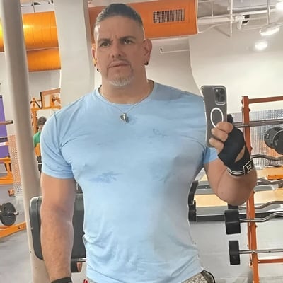 一位肌肉发达的男子在健身房自拍，穿着湿透的浅蓝色T恤，表情严肃，手持智能手机。背景中可以看到健身器材和橙色的管道。
