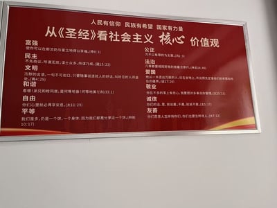 这是一块红色背景的悬挂式宣传板，顶部标注“人民有信仰 民族有希望 国家有力量”，核心主题为“从《圣经》看社会主义核心价值观”。宣传板将社会主义核心价值观的12项内容（富强、民主、文明、和谐、自由、平等、公正、法治、爱国、敬业、诚信、友善），分别对应《圣经》中的相关经文，采用左右两列的排版方式清晰展示，试图用宗教文本解读主流价值观，背景中还带有东方明珠等城市建筑剪影。