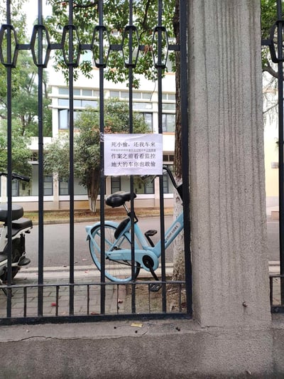 一张校园内的照片，黑色铁栅栏上挂着一张手写标语，内容为'死小偷，还我车来 作案之前看看监控 地大的车你也敢偷'，表达了自行车被盗后车主的愤怒与警告。栅栏旁停放着一辆浅蓝色自行车，背景可见校园建筑和树木，整体场景反映了校园自行车盗窃问题及车主的无奈反击。