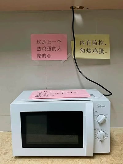 图片展示了一个办公室或共享厨房场景中的白色美的(Midea)微波炉。微波炉上方的瓷砖墙上贴着两张手写告示：粉色告示写着“这是上一个热鸡蛋的人赔的”并配有一个圆圈符号，黄色告示则写着“内有监控，勿热鸡蛋”。微波炉顶部还放着一张相同内容的粉色告示。这张图片以幽默的方式展示了因有人在微波炉中加热鸡蛋导致损坏后，其他使用者采取的警示措施和赔偿证据。