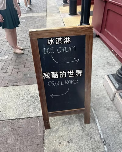 一张拍摄于城市人行道的趣味指示牌照片。画面中央是一块木质边框的黑色小黑板，上方用白色粉笔写着“冰淇淋 ICE CREAM”并配有一个向下的箭头，下方则写着“残酷的世界 CRUEL WORLD”并配有一个向上的箭头，形成有趣的文字游戏。背景可见红砖铺就的路面、穿着绿色裙子的路人腿部以及红色墙面的建筑，整体呈现出轻松幽默的街头氛围。

图片通过将甜美诱惑的“冰淇淋”与沉重现实的“残酷世界”形成鲜明对比，创造出一种黑色幽默的反差萌效果。