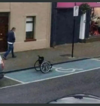 这是一张街头实拍的搞笑梗图，画面中蓝色的无障碍停车位上翻倒着一辆轮椅，旁边人行道上一名男子正迈步离开，巧合的画面看起来像是男子为了占用无障碍车位，假装成残疾人，留下轮椅就潇洒走开，利用轮椅与车位标识的位置制造出极具荒诞感的视觉效果，用来调侃占用无障碍车位的不文明行为，充满讽刺与搞笑意味。