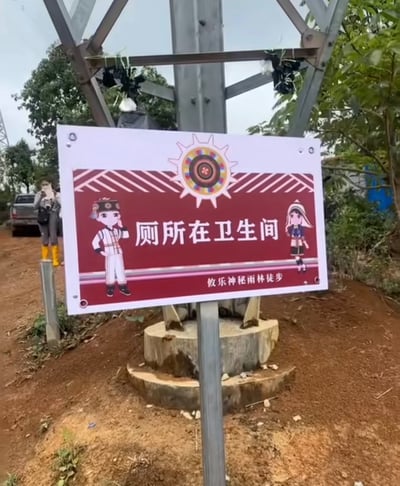这是一张在户外徒步区域拍摄的实拍照片，画面主体是立在土路边的攸乐神秘雨林徒步路线指示牌，指示牌以酒红色为主色调，搭配白色边框，顶部带有民族风格的太阳形纹饰，左右两侧分别绘制了身着少数民族服饰的男女卡通人物形象，牌面正中写着“厕所在卫生间”的指引文字，下方标注有“攸乐神秘雨林徒步”字样。该指引内容属于没有实际方位信息的同义反复，完全无法为寻找卫生间的游客提供有效帮助，反而会让询问卫生间位置的人更加困惑，非常有笑点。背景可见土路、金属电线杆、周边绿植，远处还有站着的游客和停放的车辆，结合标题“那请问卫生间在哪里”，进一步凸显了该指示牌的无厘头属性，面对该指引确实无法获知卫生间的具体方位。