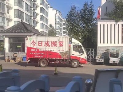 这是一张在小区出入口拍摄的日常场景照片，画面主体是一辆红白配色的「今日成搬家」厢式货车，停在小区岗亭旁。货车车身上清晰标注了可提供居民搬家、单位搬迁、设备搬运、钢琴搬运等服务，同时印有联系电话。背景可见高层居民住宅楼、中式风格岗亭、带有国旗的建筑，旁边还停放着电动三轮车，整体氛围是平实的城市社区日常，推测是搬家车辆正在开展服务或等待作业。