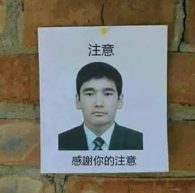 这张图是被张贴在红砖墙上的打印告示类图片，告示整体为白底，顶部印有黑色“注意”二字，中间位置是一名身穿深色西装、浅色衬衫并搭配条纹领带的东亚男性的证件照，该男性表情严肃、面无表情直视前方，告示底部印有黑色“感谢你的注意”字样，整体排版模仿警示公告、寻人启事的格式，但内容没有实际的通知或警示含义，形成荒诞搞笑的无厘头效果，常被用作网络互动中吸引他人注意力后表达感谢的梗图。