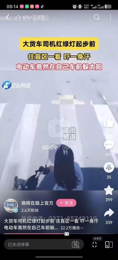 这是一段道路交通安全新闻的视频截图，画面显示在路口斑马线处，一名电动车骑手为躲避烈日，将车停在大货车的车头盲区位置；大货车司机在红绿灯起步前检查盲区，发现该情况后吓出一身冷汗。这个场景存在极大安全隐患，若货车未检查盲区直接起步，极易引发严重交通事故。图中无谐音梗、双关或暗示内容。
