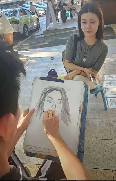 这是一张街头艺术家为女生素描画像的照片，取景于城市步行街或户外公共空间。女生坐在小板凳上，摆出自然姿势，画师专注地在画板上绘制女生的素描像。画板上已经画出头部结构和发型，现场环境活跃，侧重昭示生活气息和街头艺术的温度。整个画面呈现出一种平静、专注、艺术与生活交融的氛围。无双关或谐音梗，也无暗示内容。