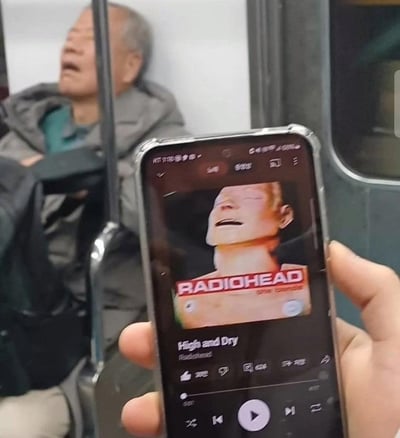 地铁场景中，一位老人仰头靠在座椅上，表情与手机屏幕上显示的Radiohead乐队《High and Dry》专辑封面人物姿态意外相似，形成有趣的巧合对比。拍摄者巧妙捕捉到这一瞬间，展现了现实生活与音乐文化的幽默碰撞。
