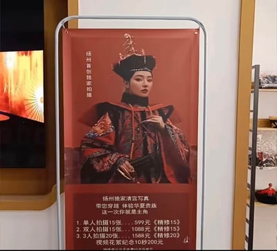 这是一款放置于线下门店的立式宣传展架，主打扬州首创独家清宫主题写真拍摄服务。展架上半部分展示了身着华丽清宫后妃服饰、佩戴精致旗头的女性模特，她妆容冷艳端庄，手持护甲，姿态优雅；下半部分则清晰列出了三种写真套餐的价格与内容，还有单独的视频花絮收费项目，搭配“带您穿越 体验华夏贵族，这一次你就是主角”的宣传语，整体风格复古庄重，旨在吸引喜爱古风、想体验清宫主题写真的消费者。
