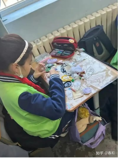 图片展示了一名身穿绿色马甲、佩戴红领巾的学生在教室书桌上DIY手工徽章或挂件，桌面上摆满了各种做工精致的徽章、织带、缝制工具和彩笔套装。包包里还塞有毛绒玩具。这反映了当下部分学生热衷于手工创作、个性表达的校园文化氛围。

未发现明显的双关或谐音梗，氛围偏日常生活趣味。