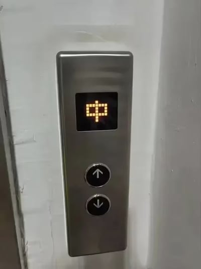 这是一张实拍的电梯外呼面板照片，银灰色金属材质的呼叫面板安装在白色墙面上，面板上方的点阵LED显示屏亮起的点阵图案恰好组合成完整的中文“中”字，下方配有向上、向下两个黑色圆形的电梯呼叫按钮。这个显示的“中”字刚好呼应河南方言里表示认可、同意含义的“中嘞”，形成了充满趣味的巧合效果。
