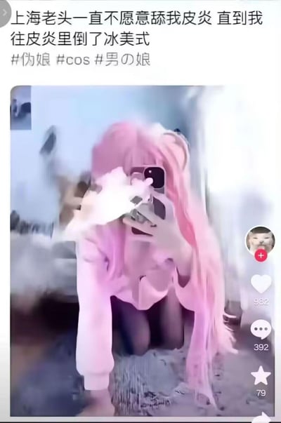这是一张竖屏短视频平台的截图，画面主体是一位留着粉色长发、身着粉色上衣的伪娘正拿着手机对镜自拍；画面上方配有低俗谐音梗文案“上海老头一直不愿意舔我皮炎 直到我往皮炎里倒了冰美式”，其中“皮炎”是“皮燕”的谐音，暗指臀部，属于擦边恶趣味内容，附带#伪娘 #cos #男の娘 三个标签。截图右侧显示了该短视频的互动数据：点赞982、评论392、收藏79，还有一个带有猫脸头像的账号关注按钮，整体是网友为博眼球创作的低俗整活内容。