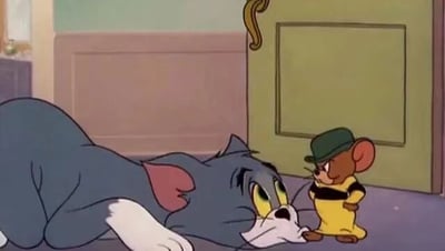这是经典动画《猫和老鼠》的截图，画面呈现了房间内的场景：蓝色的汤姆猫趴在地板上，下巴贴地，眼神低垂，神态看起来低落又带着讨好感；一旁的杰瑞鼠戴着深绿色帽子，穿着黄黑配色的制服，双手叉腰站在汤姆面前，表情严肃，姿态颇具气势，二者的姿态形成鲜明反差，背景是房间的门与浅色调的墙面。