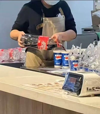 这是一张在快餐店柜台拍摄的真实场景照片，画面中一位身着黑色上衣、灰色围裙并佩戴口罩的工作人员，正手持一瓶可口可乐，将瓶中的可乐倒入印有百事可乐品牌标识的纸杯中。柜台台面上还摆放着多个同款百事可乐纸杯、包装好的餐品以及收银设备，背景是快餐店的操作区域。该场景呈现出可口可乐与百事可乐这两大竞品品牌的有趣反差，用竞品饮品来填充自家品牌容器的操作显得十分滑稽，充满了生活中的趣味感。