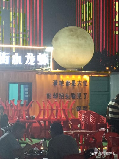 夜晚的保定户外小龙虾大排档，场地摆放着红色塑料桌椅，数位顾客正在桌前用餐。店铺上方设有“街小龙虾”的发光招牌，还有一个大型仿真月球装置，背景是红黄色竖条组成的灯光装饰。店铺墙面上印着“满地都是大便，他却抬头看见了”的文案，搭配上方的月球，营造出在接地气的市井烟火中仰望浪漫的强烈反差感，用直白的文案传递出在琐碎生活里追寻美好的意境。