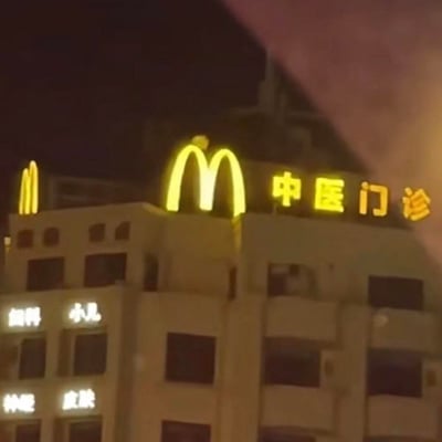 这是一张夜晚拍摄的建筑实拍图，画面中右侧建筑顶部的灯光标识，巧合地将麦当劳的金色「M」标志与「中医门诊」字样相邻排列，再加上左侧建筑也有一个类似M的灯光标识，形成了仿佛「麦当劳开中医门诊」的搞笑视觉错位效果。建筑下方还标注了「妇科、小儿、神经、皮肤」等门诊科室字样，这种品牌与医疗门诊的违和组合，充满了滑稽感，让人忍俊不禁。