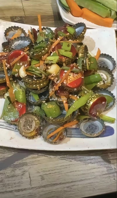 一张创意料理照片，展示了一盘由啤酒瓶盖和蔬菜混合而成的奇特凉拌菜。盘子里装满了各种颜色的啤酒瓶盖，有红色、金色、银色等，混合着绿色的青椒条、橙色的胡萝卜丝和白色的芝麻。背景中可以看到另一个装有胡萝卜和黄瓜条的盘子。这道菜的创意在于将通常会被丢弃的啤酒瓶盖作为食材容器或装饰，与新鲜蔬菜拌在一起，形成视觉冲击力强的料理。