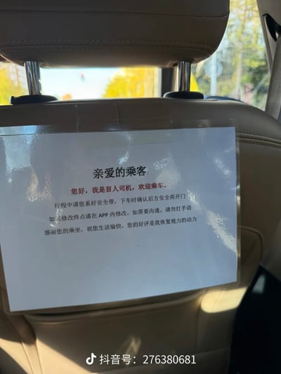 这是一张实拍的网约车内部照片，拍摄视角为车辆后排乘客位。浅米色的汽车前排座椅靠背后侧，贴有一张过塑的纸质提示告示，告示内容玩梗自称是盲人司机，依次告知乘客乘车需系安全带、下车确认后方安全、修改终点在APP操作、沟通不要打手语，最后还调侃乘客的好评是自己恢复视力的动力。图片底部带有抖音账号水印，告示内容为搞笑玩梗性质，并非真的由视障人士驾驶车辆。
