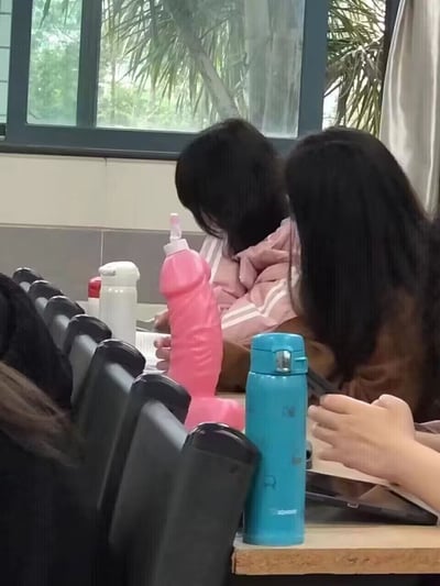 这是一张室内教室的实拍静态图，拍摄场景为阶梯式教室，前景是排列的黑色座椅靠背，前方课桌上摆放着三个水杯：最显眼的是一个粉色塑料水杯，外观造型较为特殊，容易引人产生趣味联想，旁边分别是蓝色弹盖保温杯、白色保温杯。桌面后坐着几名学生，仅露出背影和部分手臂，背景是教室的玻璃窗，窗外可见棕榈树等绿植。