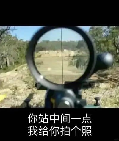 这是一张网络流行的搞笑梗图，采用第一人称视角呈现，画面主体是狙击枪的瞄准镜视野，十字准星对准了野外开阔的户外场地，瞄准镜外可见周边的树木、荒地等野外环境，图片底部黑色区域配白色文字“你站中间一点 我给你拍个照”，将用枪械瞄准锁定对方的危险行为包装成要给对方拍照的友好行为，利用瞄准镜十字和相机取景框视觉相似的特点制造反差笑点。