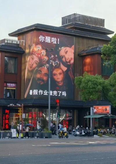 一张街景照片，展示了一栋具有传统中式风格的建筑，其外墙上竖立着一块大型垂直广告牌。广告牌上印有《西游记》师徒四人（孙悟空、唐僧、猪八戒、沙和尚）的特写肖像，人物表情夸张。广告牌顶部文字为“你醒啦！”，底部文字则是灵魂拷问“暑假作业做完了吗”，通过经典文学角色与现代学生暑期焦虑的结合制造幽默效果。建筑周围有行人和停放的电动车，呈现出日常街景氛围。