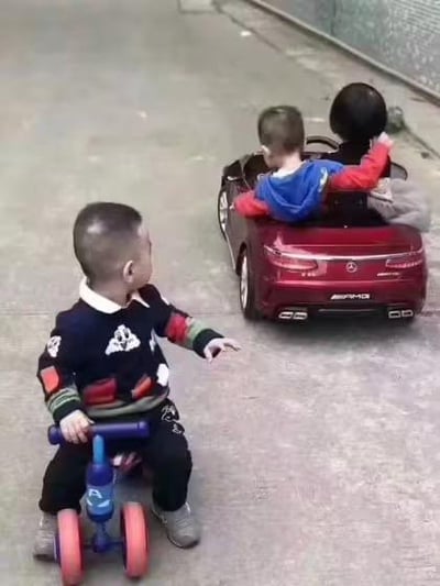 画面里有三个小朋友，两个坐在一辆红色奔驰样式的电动玩具车内，后座的小朋友回头看向后方的伙伴，还伸出一只手；另一个小朋友坐在一辆简易的蓝色三轮脚踏玩具车上，伸手指向前面的电动玩具车，眼神满是羡慕。这个场景生动还原了现实里不同的出行方式差距，充满搞笑又真实的反差感，常被用来调侃现实中的贫富差距类话题。