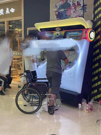 在商场的公共区域，一位原本乘坐轮椅的玩家正站在一台类似maimai的节奏音游机前专注操作游玩，他的轮椅停放在身旁。周围还有其他路人，背景可见一家名为“煎点心”的中式点心餐厅，整体场景传递出玩家对游戏的热爱，展现了打破身体限制的积极状态。
注：图中没有双关、谐音梗或暗示类内容。