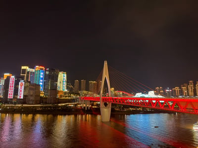 这是一张夜晚拍摄的重庆城市江景实拍图，画面中心是一座带有白色异形桥塔的斜拉桥，桥身被红色灯光照亮十分醒目，斜拉索呈暖橙色。桥下是宽阔的江面，水面清晰倒映着大桥和岸边楼宇的各色灯光，色彩斑斓。背景是林立的重庆CBD高层建筑，楼体亮着商业广告、企业标识与宣传类灯光文字，远处还能看到红色拱形桥梁和造型独特的公共文化建筑，整体灯火璀璨，展现出重庆山城特有的繁华夜景观感。