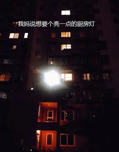这是一张夜晚拍摄的居民楼实拍静态图，画面中整栋居民楼整体处于昏暗的夜色中，只有少数住户的窗户透出正常的室内灯光，唯独中间某楼层的一户住户的厨房位置安装的灯亮度过高，光线强烈到过曝发白，在昏暗的楼体中格外突兀显眼。图片上方配有文字“我妈说想要个亮一点的厨房灯”，用现实中远超预期的夸张亮度形成反差搞笑效果，呼应标题“谁掐的瞬爆”的玩梗调侃，将过亮的灯光调侃成爆炸产生的强光。
