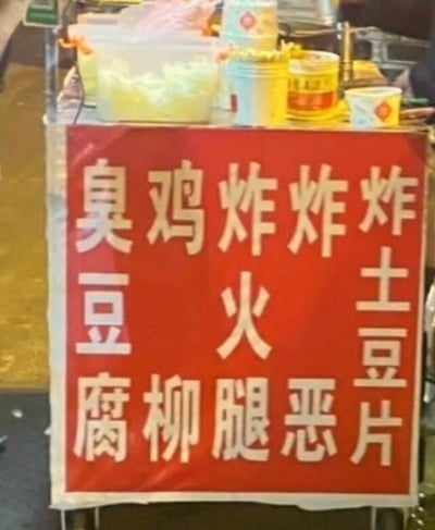 这是一张实拍的街边小吃摊场景图，摊位前方悬挂着红底白字的售卖品类招牌，因摊主排版失误，原本应该竖排读取的售卖品类（臭豆腐、鸡柳、炸火腿、相关炸物、炸土豆片），横排阅读时会出现“臭鸡炸炸炸”“腐柳腿恶片”等莫名其妙的内容，十分滑稽好笑。招牌后方可见摊位上摆放的塑料储物盒、调料罐、一次性纸杯等小吃经营常用物品，整体是夜间街边摆摊的日常场景。