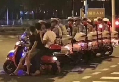 这是一张夜晚马路场景的真实拍摄图，画面左侧一辆红色电动车违规搭载了三名人员，画面右侧的一整排交警骑着带警灯的摩托车，从拍摄角度看仿佛正紧紧跟在电动车后方，营造出电动车违规被交警队伍盯上的紧张氛围。但实际上这是典型的视觉错位效果，交警队伍与电动车处于不同车道，并非在追踪该电动车，只是拍摄视角让两者在画面中重叠，形成了极具戏剧性的乌龙搞笑场景。