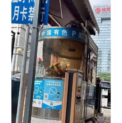 这是一张户外实拍的静态图片，主体是一个带有“中国有色 P”标识的停车场岗亭，岗亭为玻璃与金属结合的结构，顶部设有蓝色的标识牌。岗亭的玻璃外侧贴有蓝色的提示海报，上面印有“一车不能占多位 违规停车要缴费”的提示语，搭配卡通图案与二维码，用于规范停车行为。岗亭旁立着一块蓝色的交通指示牌，标注“月卡禁行”。岗亭内部可见摆放的花束，背景是现代化的高楼建筑，其中一栋楼体上带有“北新”的品牌标识，整体场景位于城市中的停车场入口区域，呈现出日常的停车管理场景，环境整洁规整。