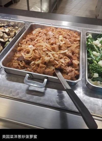 这是一张食堂取餐区的实拍图，三个不锈钢餐盆依次摆放，左侧是烧茄子类菜品，中间餐盆内是带汤汁的洋葱炖肉块，右侧是西兰花等清炒时蔬。但图片底部标注的文字却是「英国菜洋葱炒菠萝蜜」，实际菜品和标注完全不符，形成强烈反差，用来吐槽食堂菜品名不副实的搞笑场景，引发对食堂奇葩菜名的共鸣。