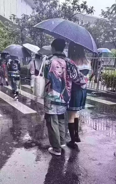 雨天的户外斑马线区域，地面因降雨变得湿漉漉的，一对男女共撑一把黑色雨伞并肩前行。男生的外套背后印着一位粉发笑容灿烂的动漫女孩，从背后视角看去，这个图案恰好和两人的位置形成了有趣的视觉错位，仿佛这个动漫女孩被夹在两人中间，制造出类似“电灯泡”的搞笑既视感，场景充满二次元趣味。