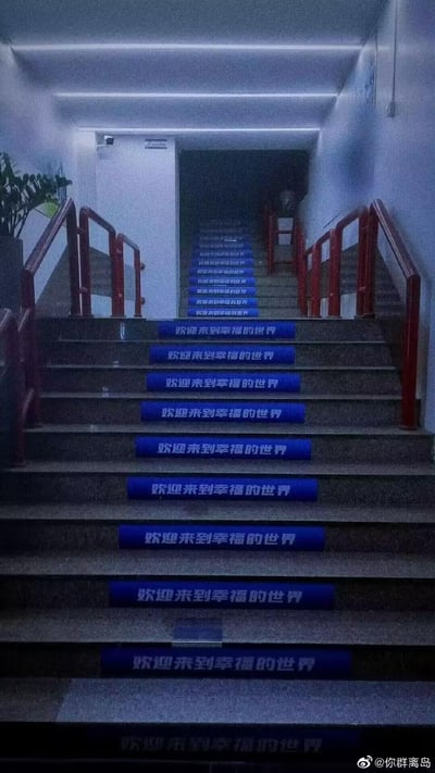 这是一张室内向下楼梯的实拍图，每一级台阶的前沿都粘贴着蓝底白字的标语贴纸，内容均为「欢迎来到幸福的世界」，重复的标语沿着楼梯一直延伸至下方，搭配红色的楼梯扶手，营造出一种沉浸式的引导感，仿佛走下楼梯就能踏入一个被定义为「幸福」的专属空间，整体氛围温馨又充满仪式感。