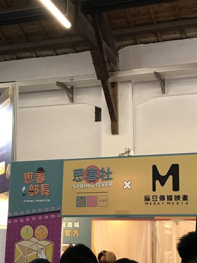 这是一张室内展会现场的实拍照片，展示了「思春部长」「思春社（SPRING FEVER）」与「麻豆傳媒映畫（Model Media）」的联合展位。展位招牌采用复古趣味的设计风格：左侧蓝底区域印有戴礼帽叼烟斗的卡通元素与「思春部长 SPRING MINISTER」字样；中间黄底区域标注「思春社 SPRING FEVER」，并以「×」连接右侧的「麻豆傳媒映畫 Model Media」黑色M标识，下方还有相关小图标。背景是带有木质横梁的厂房式白色墙面，画面下方能看到部分观众的头顶，显示出现场有一定人流量。