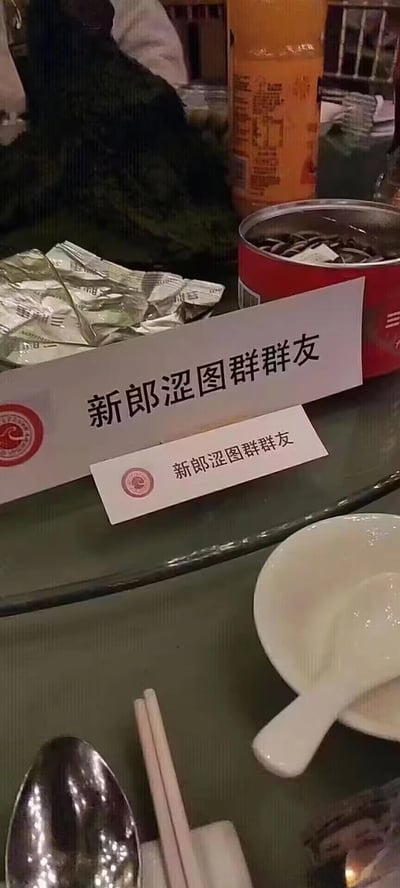 这是一张婚礼宴席餐桌上的实拍图，桌面摆放着一大一小两个席位卡，上面都印着「新郎涩图群群友」的字样，旁边还有罐装食品、小包装零食和餐具，整体是典型的中式婚礼聚餐场景。
这是朋友间的玩梗操作，「涩图群」是网络上指代分享擦边趣味图片群组的说法，将这个名称用在正式的婚礼席位卡上，形成强烈的反差感，充满搞笑滑稽的氛围，体现了新郎和朋友们之间的轻松玩笑关系。